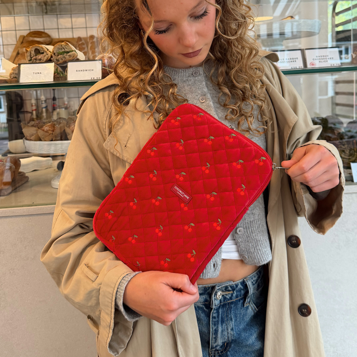 Woman in a tan coat holding a quilted cherry-red velvet iPad sleeve near a bakery display case.