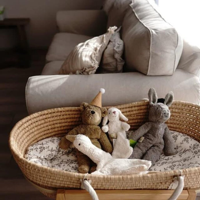 Wicker basket with stuffed animals includeing a Senger beige teddy bear on a floral-patterned cushion in front of a beige sofa.