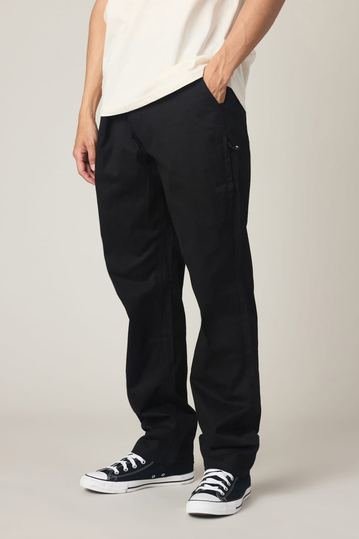 A person stands with one hand in their pocket, wearing a white t-shirt, the 686 Men's Everywhere® Standard Pant - Relaxed Fit by 686 in black, and black Converse sneakers. The photo is cropped from the shoulders down.