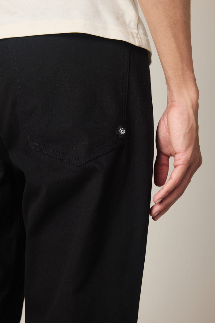 A person wearing 686 Men's Everywhere® Standard Pant - Relaxed Fit in black and a light shirt is shown from the waist down, with their right hand resting near the back pocket.