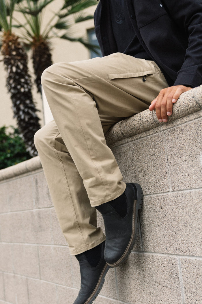 A person wearing 686 Men's Everywhere® Standard Pant - Relaxed Fit in tan and black boots sits on a stone ledge, with palm trees and a building in the background. Only the lower half of their body is visible.
