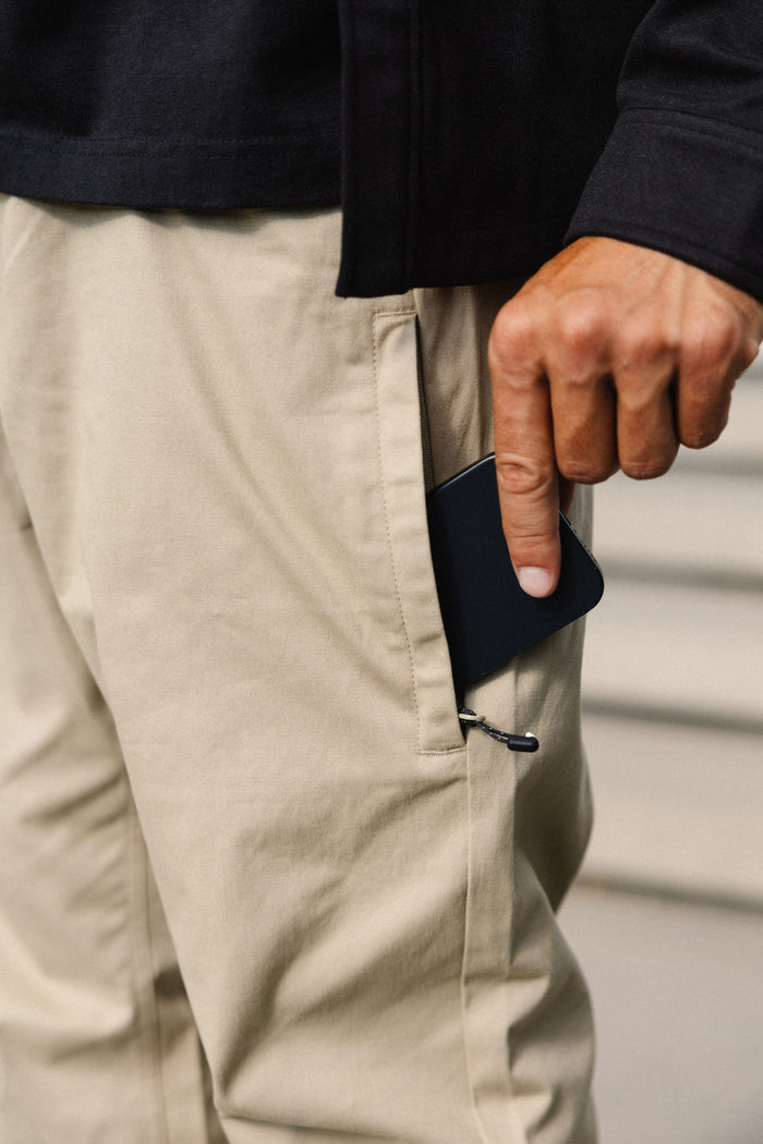 A person wearing the 686 Men's Everywhere® Standard Pant - Relaxed Fit in beige is placing a black smartphone into the zippered side pocket with their right hand. Only the hand, pocket, and part of the pants and jacket are visible.