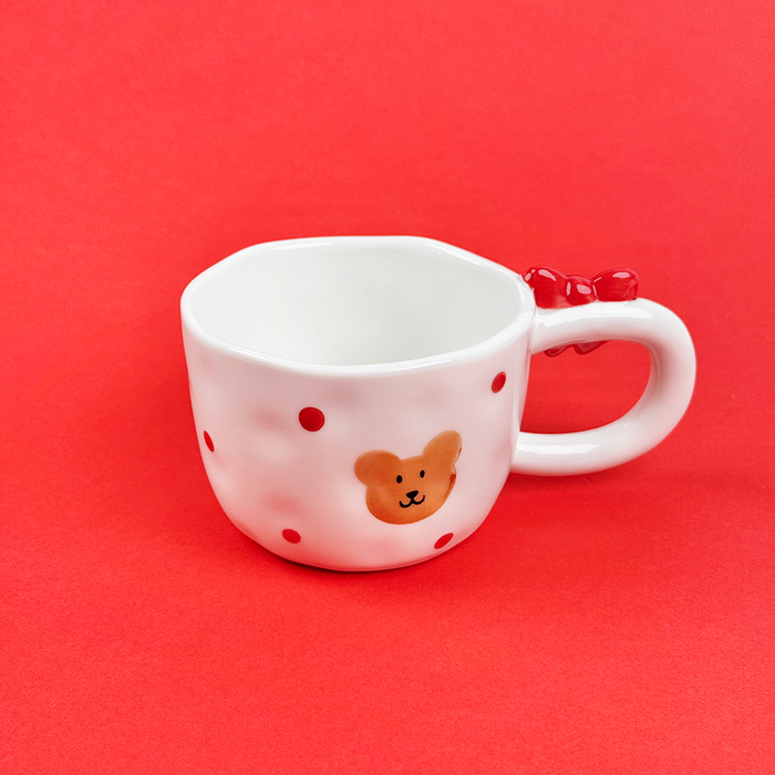 White mug with red dots, bear face, and a bow-shaped handle, shown on a red background.