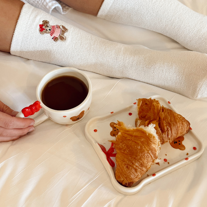 Person in bed with bear-print socks holds a pink Beary Cute mug and toast-shaped plate next to croissants.