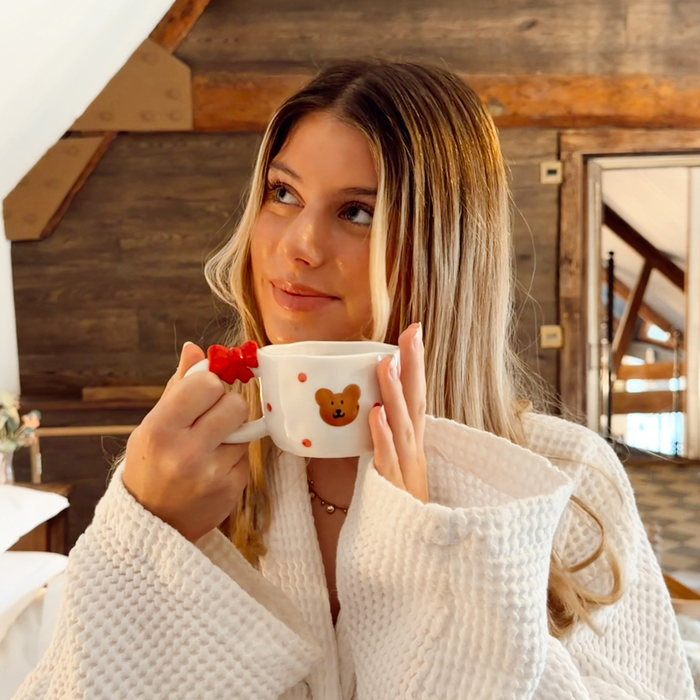 Woman in a white robe holds Coconut Lane’s Beary Cute ceramic mug and toast plate set in a wooden room, smiling.