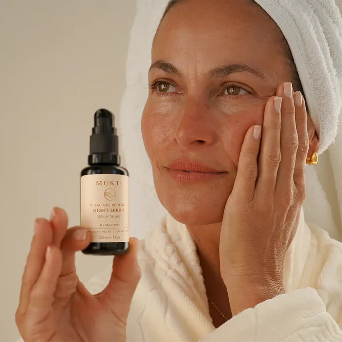 A woman in a white robe and towel cap holds a small black bottle while touching her cheek.