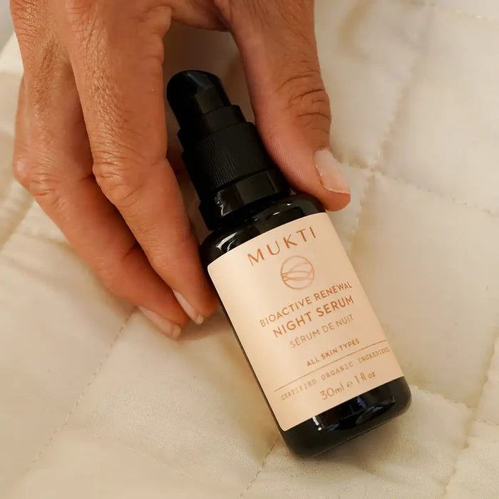 A hand holds a small black pump bottle of MUKTI Bioactive Renewal Night Serum against a soft white quilted fabric background.