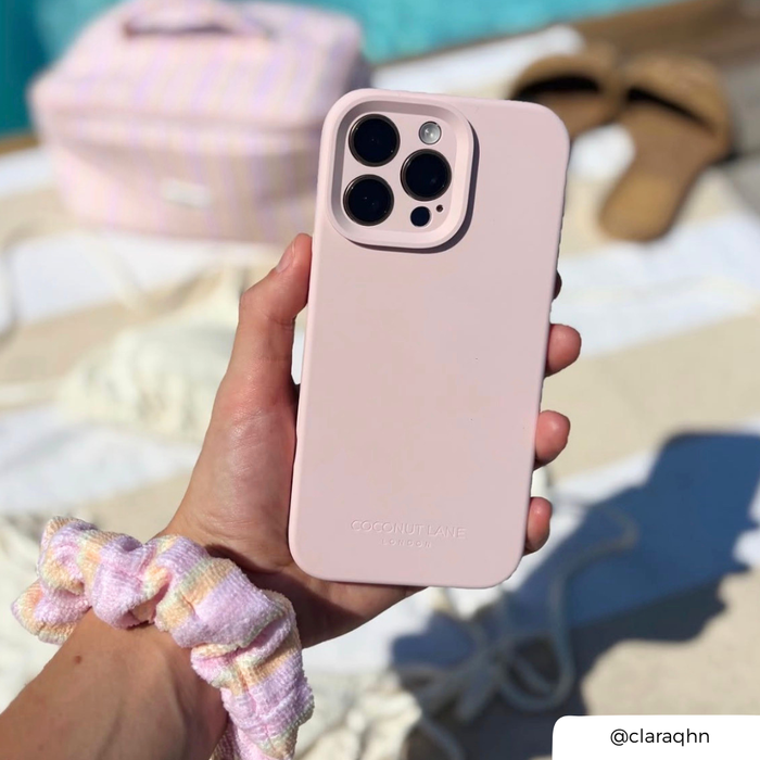 A hand holds a light pink phone case with a raised camera cutout, shown against a softly blurred beach-like background with a striped bag, sandals, and a blue towel. The case has subtle branding near the bottom.