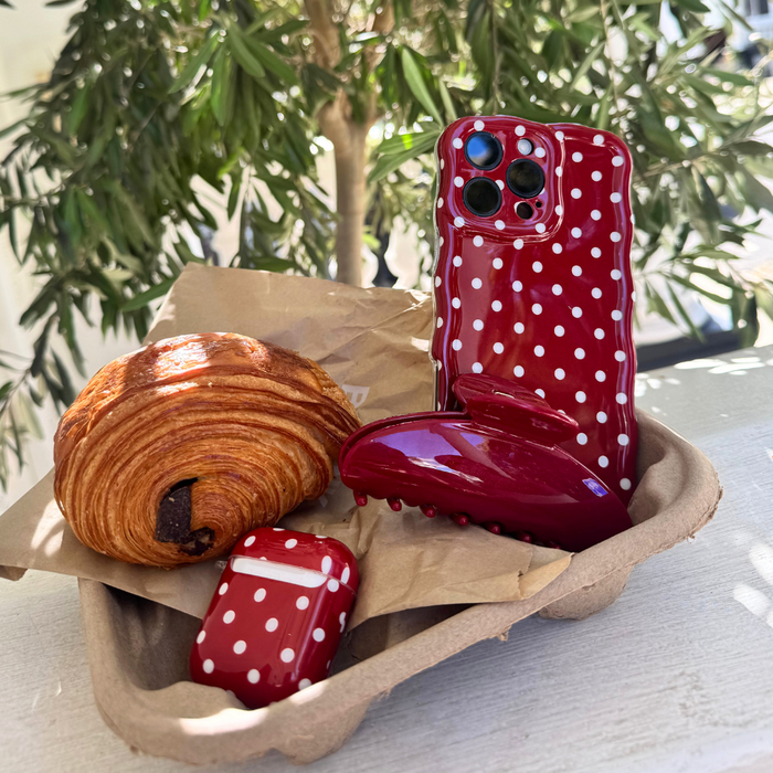 Cherry red claw clip, matching phone and AirPods cases, and a croissant on a tray with leafy plant behind.