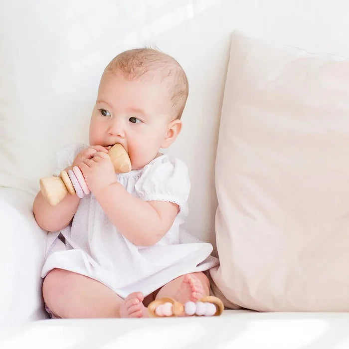 Baby in a white onesie holding a wooden baby rattle on a white couch.