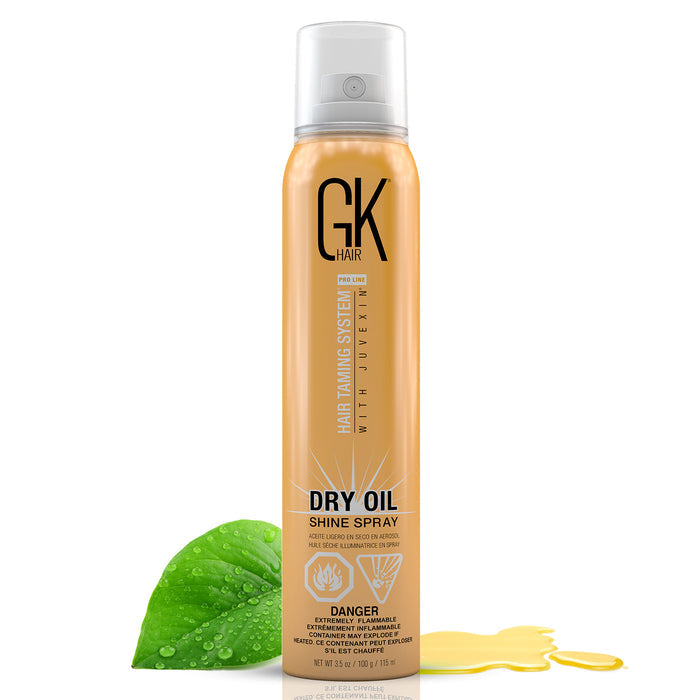 GK Hair Dry Oil Shine Spray 115 ml gold aerosol can with a clear cap, beside a dewy green leaf and a pool of golden oil