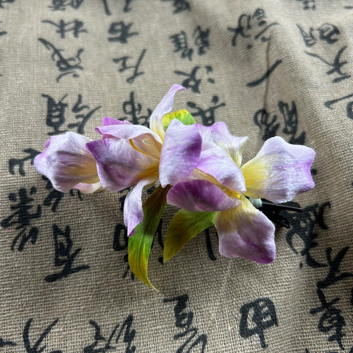 Handmade purple Ronghua silk iris flower hair clip displayed on traditional Chinese calligraphy fabric background