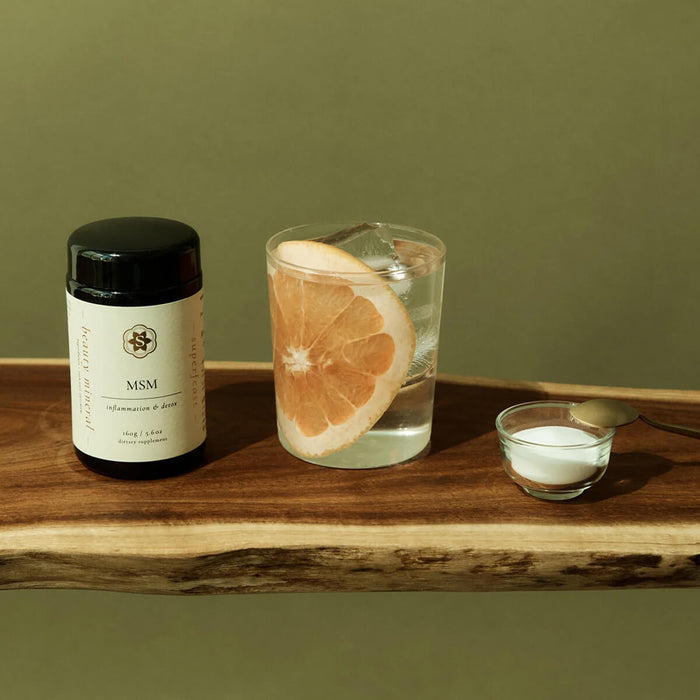 MSM supplement bottle, glass of water with a slice of orange, and small bowl on a wooden surface with a green background.