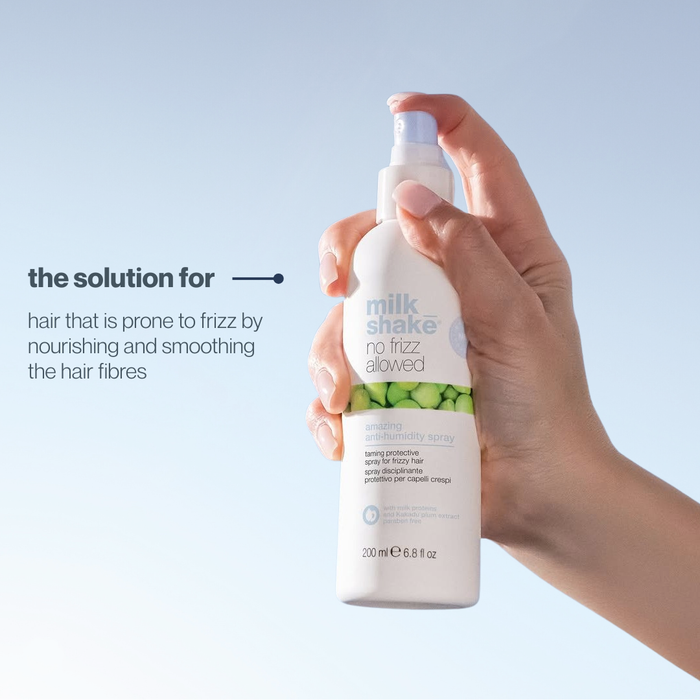 Hand holding a bottle of 'milk_shake' no frizz allowed amazing anti-humidity spray against a light blue background