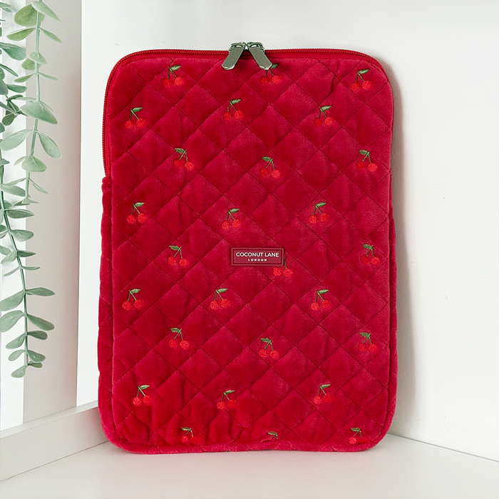 Red quilted velvet iPad sleeve with small cherry pattern, standing upright next to a green plant.