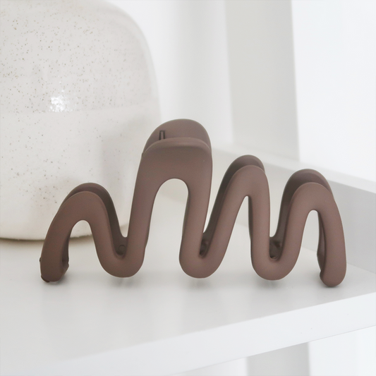Mocha-colored squiggle claw clip on a white shelf next to a round, speckled ceramic piece.