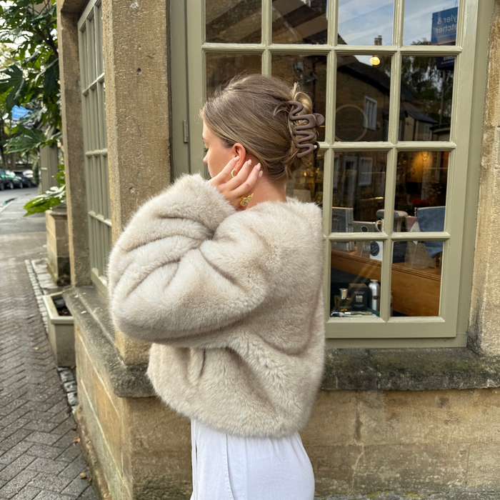Woman in beige faux fur coat wears Coconut Lane Mocha Squiggle Claw Clip, standing by large windows.