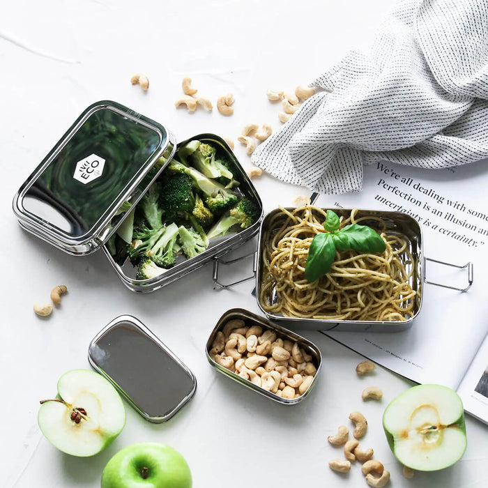 Three stainless steel bento boxes packed with broccoli and spaghetti, placed on a white surface with scattered fruit and nuts.