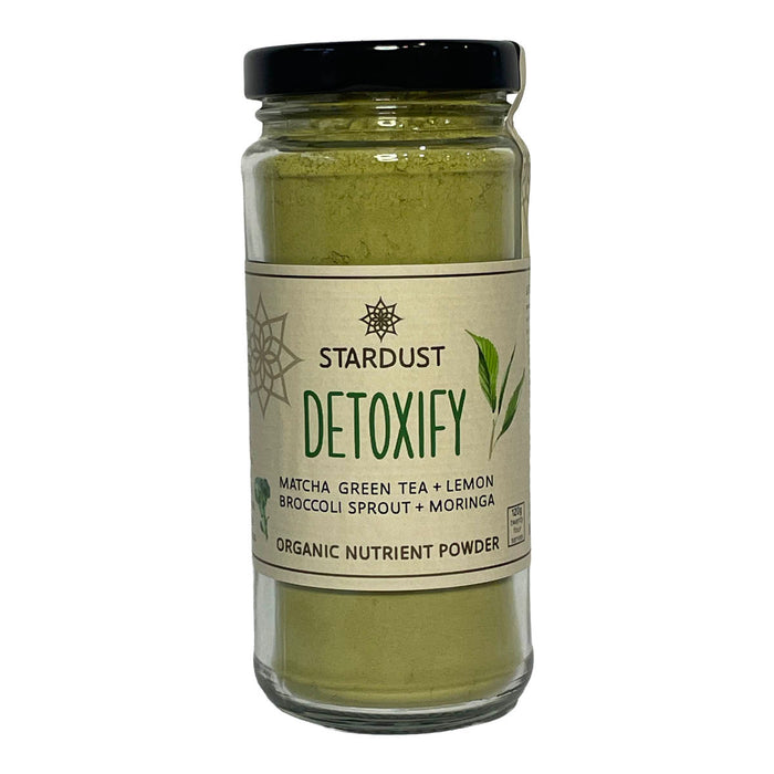 A glass jar of Stardust Detoxify organic nutrient powder, filled with a light green powder and topped with a black lid, set against a plain white background.