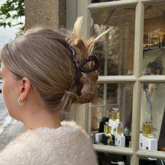 Woman with blonde hair wearing a chocolate Coconut Lane wavy claw clip, gold hoops, and a fuzzy coat by a window.